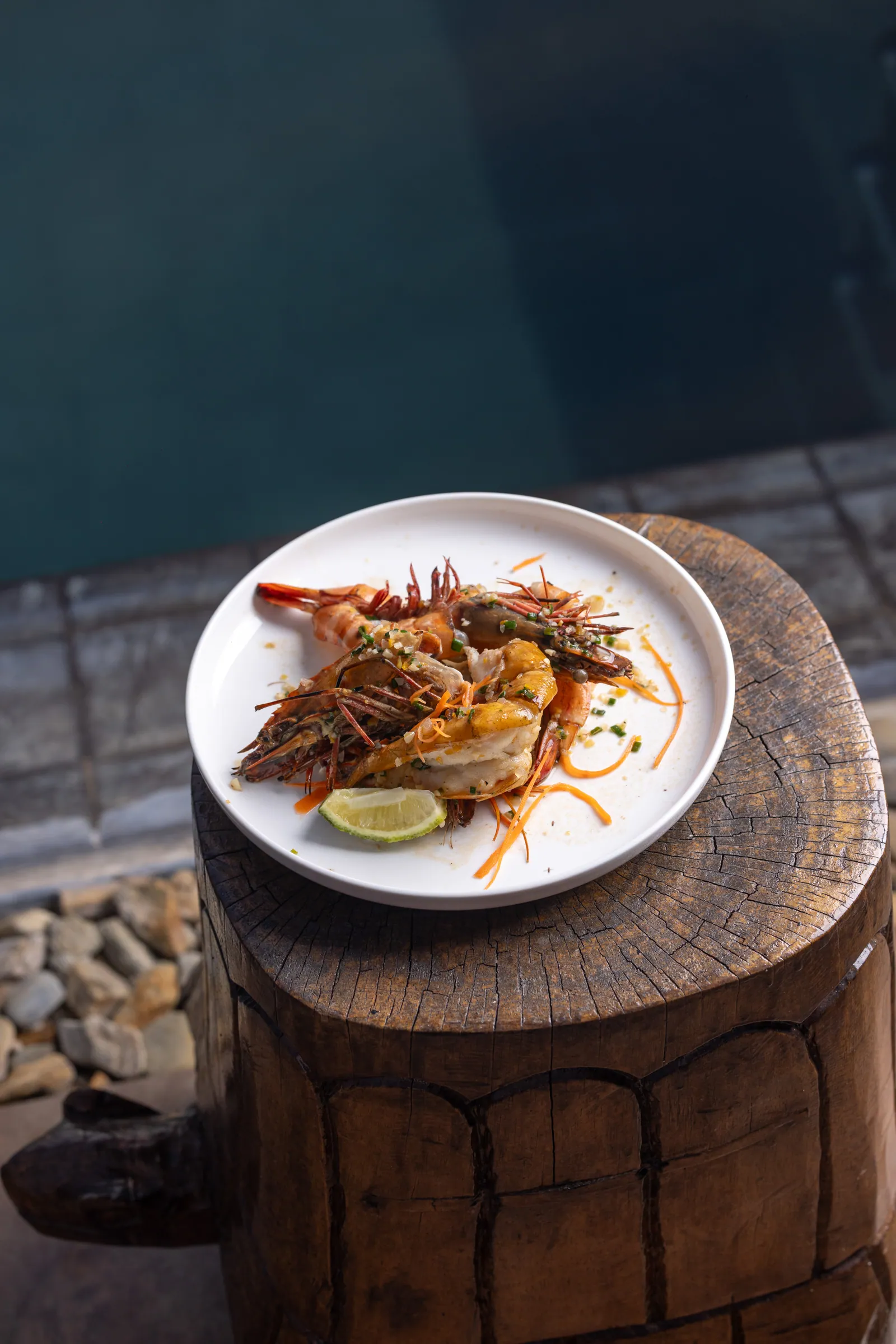 Grilled prawns and lime on a white plate, set on a carved wooden block by the pool